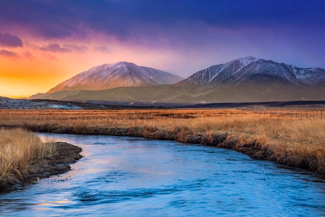 Sunrise over a serene river with snow-capped mountains in the background and golden grasslands in the foreground, capturing the tranquil beauty of Bishop, California in the heart of the Eastern Sierra. visit bishop