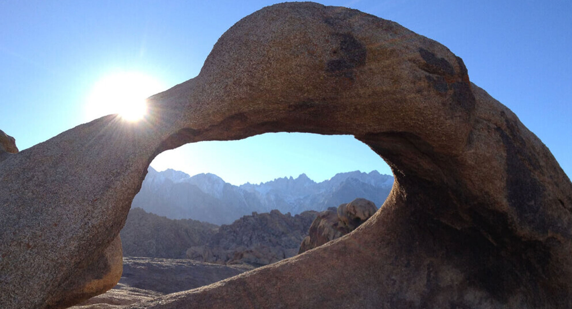 Sun shining through a rock arch in Bishop, California, with the majestic Eastern Sierra mountains and a clear blue sky in the background. visit bishop