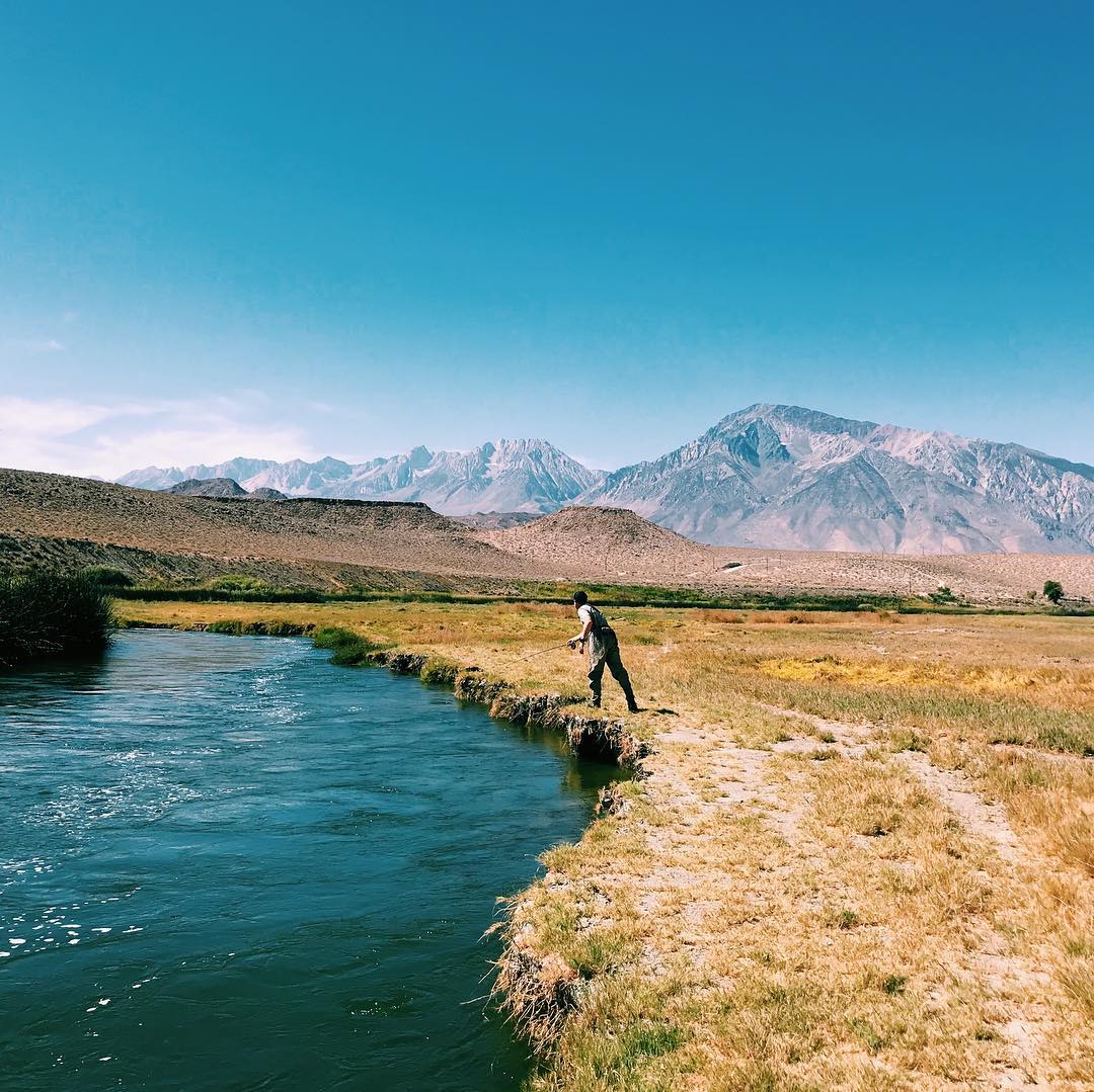 Person fishing by a river in Bishop, California, with mountains of the Eastern Sierra in the background under a clear blue sky. visit bishop