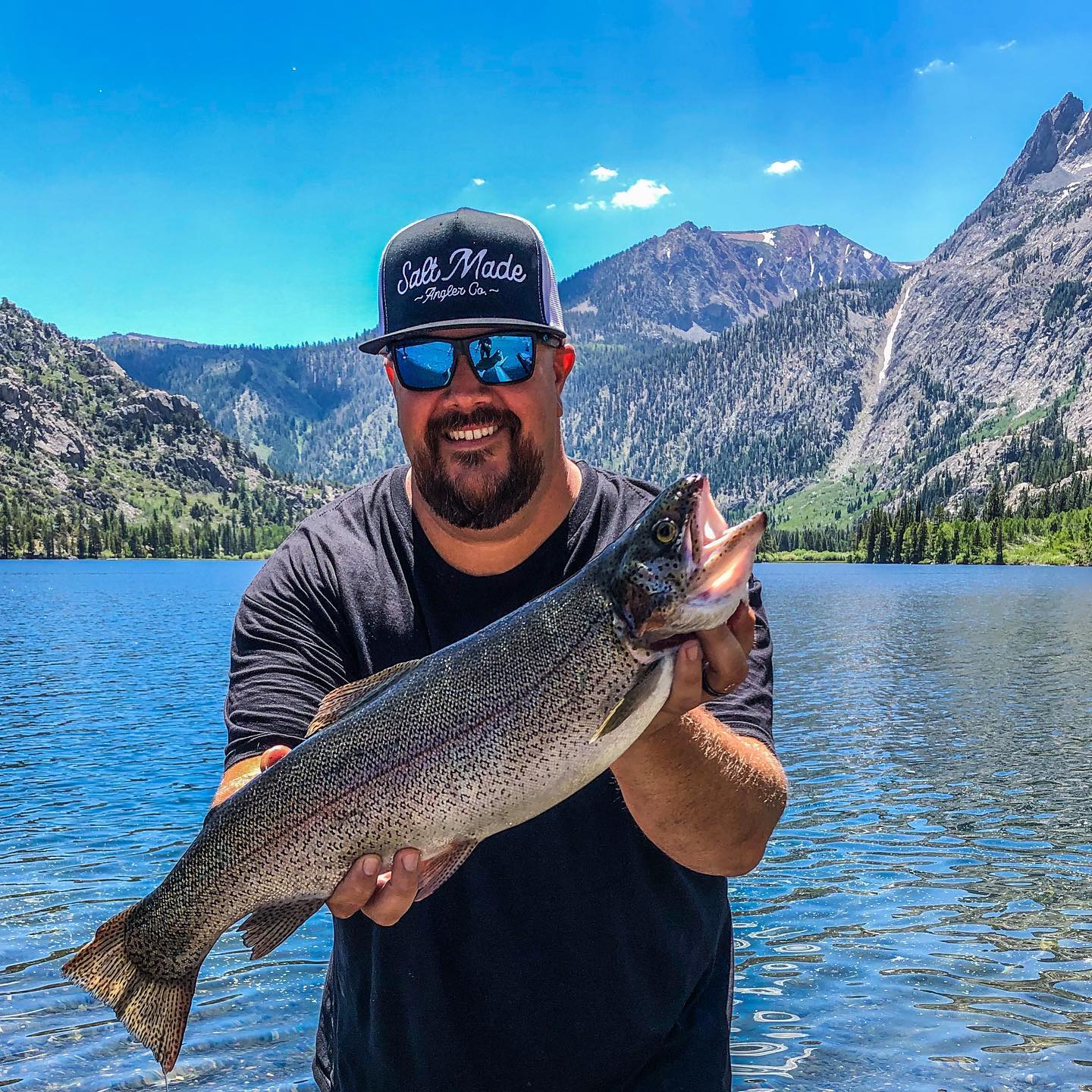 Man wearing sunglasses and a cap, smiling and holding a large fish in front of a scenic lake and mountain landscape in Bishop, California. visit bishop