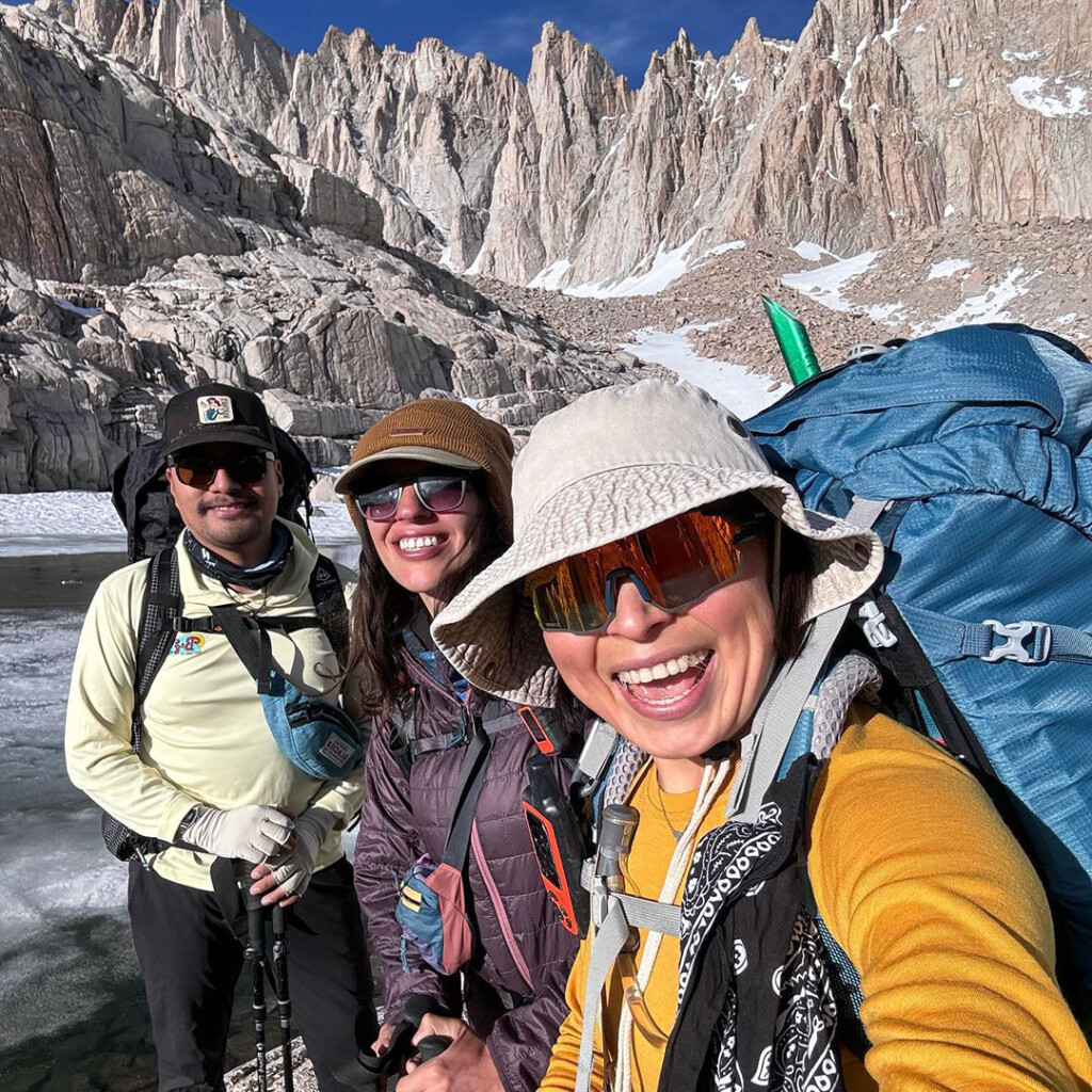 Three hikers, with backpacks and sunglasses, smiling in front of the breathtaking Bishop California landscape in the Eastern Sierra. visit bishop