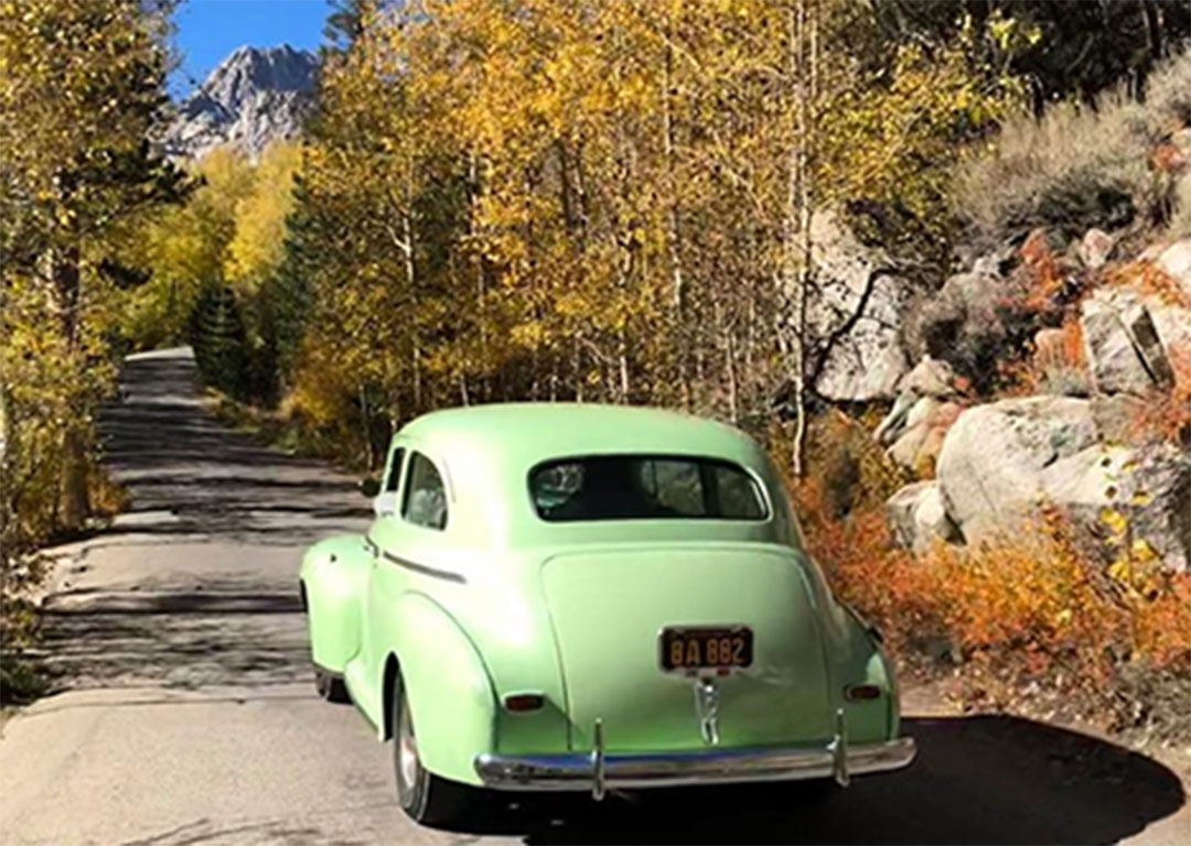 A vintage green car drives down a winding road surrounded by autumn trees and the distant mountains of the Eastern Sierra near Bishop, California. visit bishop