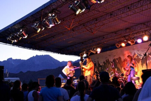 A band performing live on an outdoor stage at dusk with a mountainous backdrop of the Eastern Sierra and an audience watching in Bishop, California. visit bishop
