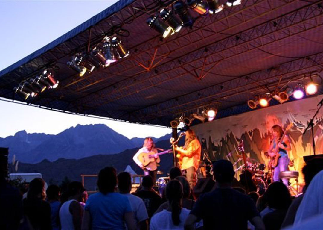 A band performing live on an outdoor stage at dusk with a mountainous backdrop of the Eastern Sierra and an audience watching in Bishop, California. visit bishop