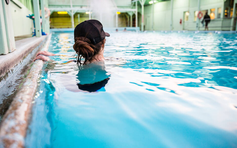 Person in a black cap relaxing in an indoor swimming pool, holding onto the pool edge, facing away from the camera, reminiscent of serene afternoons in Bishop, California. visit bishop