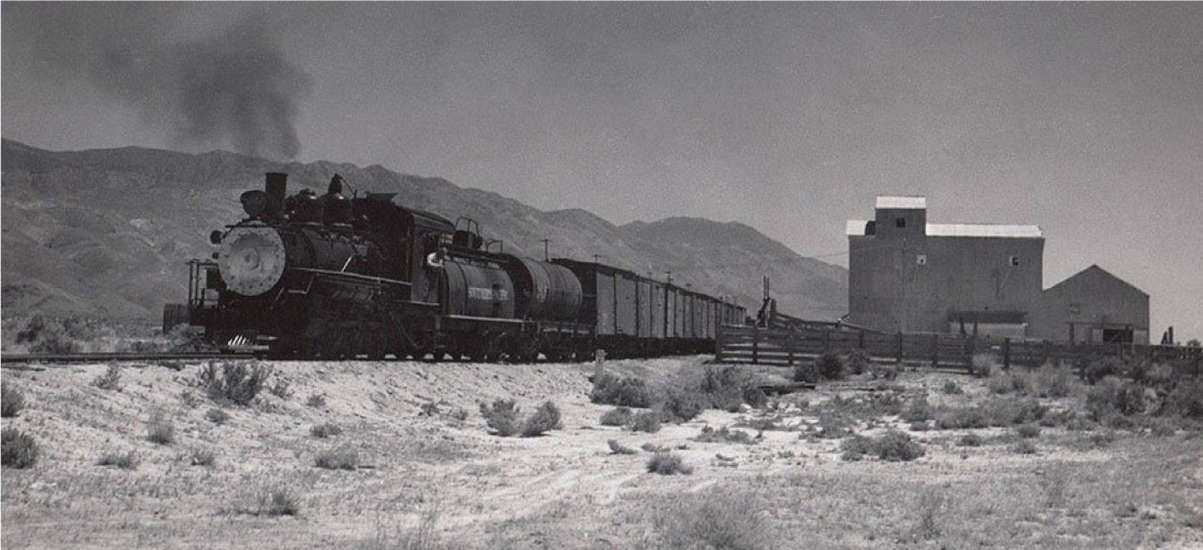 A vintage steam locomotive pulls a freight train through the desert landscape of Bishop, California, with the Eastern Sierra mountains and historic buildings in the background. visit bishop