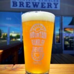 A glass of beer with the "Mountain Rambler Brewery" logo sits on a wooden table outside the brewery in Bishop, California, framed by the stunning backdrop of the Eastern Sierra. visit bishop