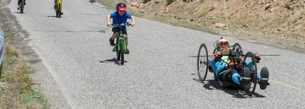 Four cyclists, one on a handcycle, ride uphill on a sunny day along a winding road in a rocky, wooded area. visit bishop