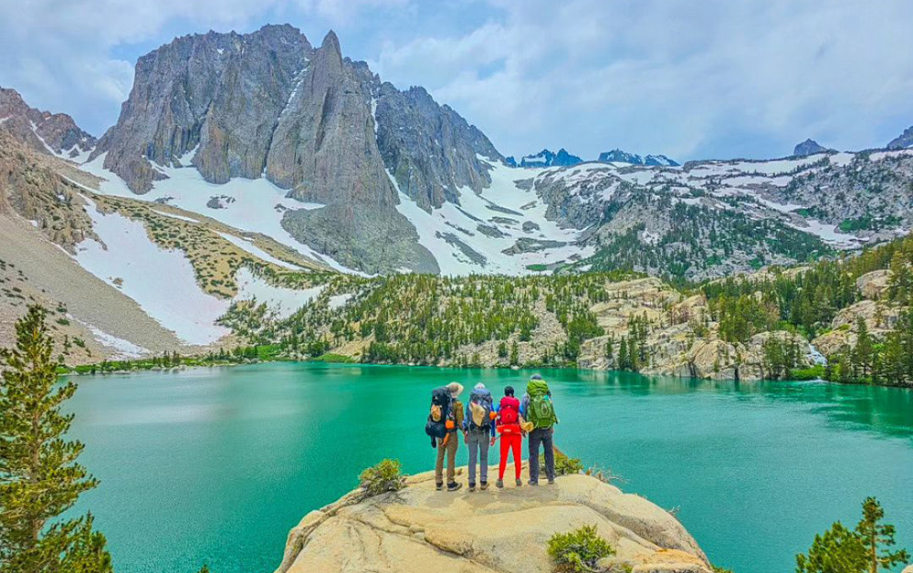 Four people wearing backpacks stand on a large boulder overlooking a turquoise lake with mountain peak in background