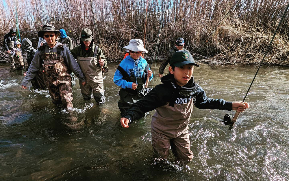 Kids carrying fishing rods, wearing waders, wading through shallow river