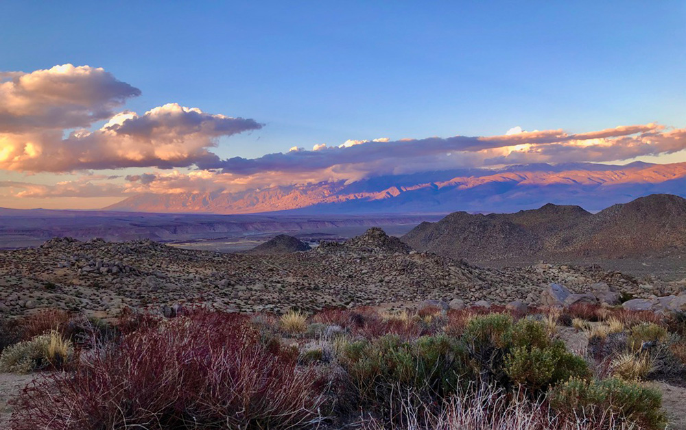 Sunset over high desert scrub, mountains in distance, clouds overhead, in orange and red glow.