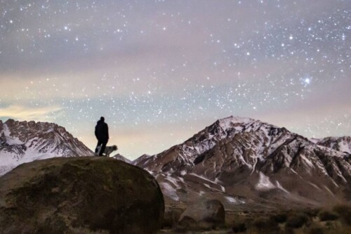 Man and dog stand on huge boulder looking at stars in evening sky, snow capped mountains form backdrop. visitbishop
