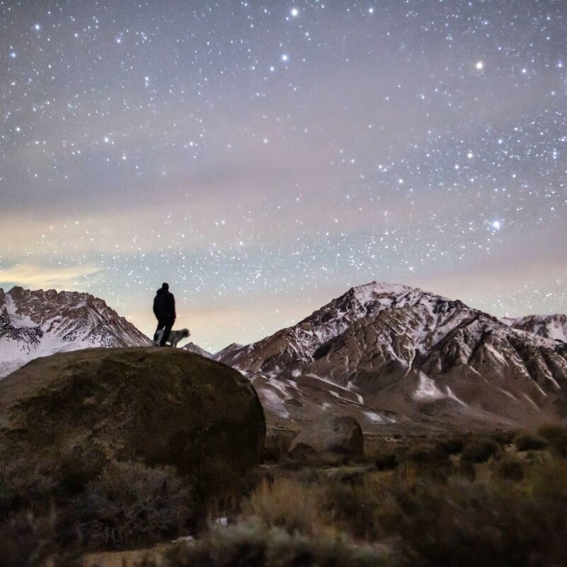 Man and dog stand on huge boulder looking at stars in evening sky, snow capped mountains form backdrop. visitbishop