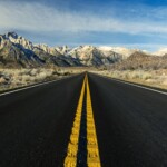 Double yellow center line of black highway with snow capped mountains in the distance.