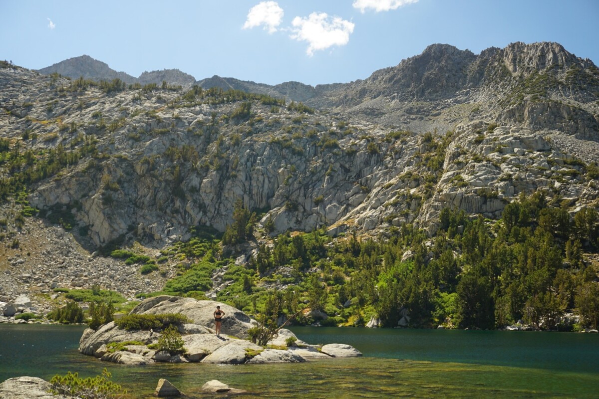 A person stands on granite boulders beside a clear emerald alpine lake, surrounded by pine trees and rugged mountain peaks near Bishop, CA