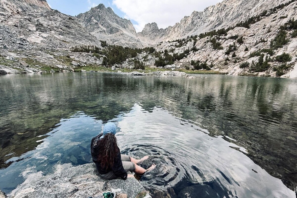 A hiker sits barefoot at the edge of a crystal-clear alpine lake, dangling feet in the water, with jagged granite peaks reflecting on the calm surface, near Bishop, CA