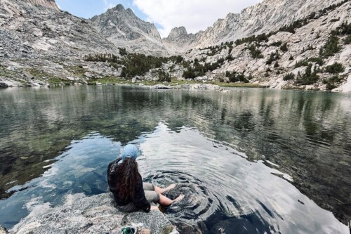 A hiker sits barefoot at the edge of a crystal-clear alpine lake, dangling feet in the water, with jagged granite peaks reflecting on the calm surface, near Bishop, CA