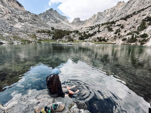A hiker sits barefoot at the edge of a crystal-clear alpine lake, dangling feet in the water, with jagged granite peaks reflecting on the calm surface, near Bishop, CA