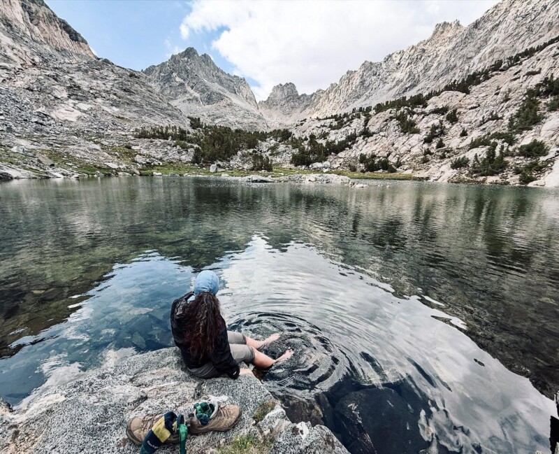A hiker sits barefoot at the edge of a crystal-clear alpine lake, dangling feet in the water, with jagged granite peaks reflecting on the calm surface, near Bishop, CA